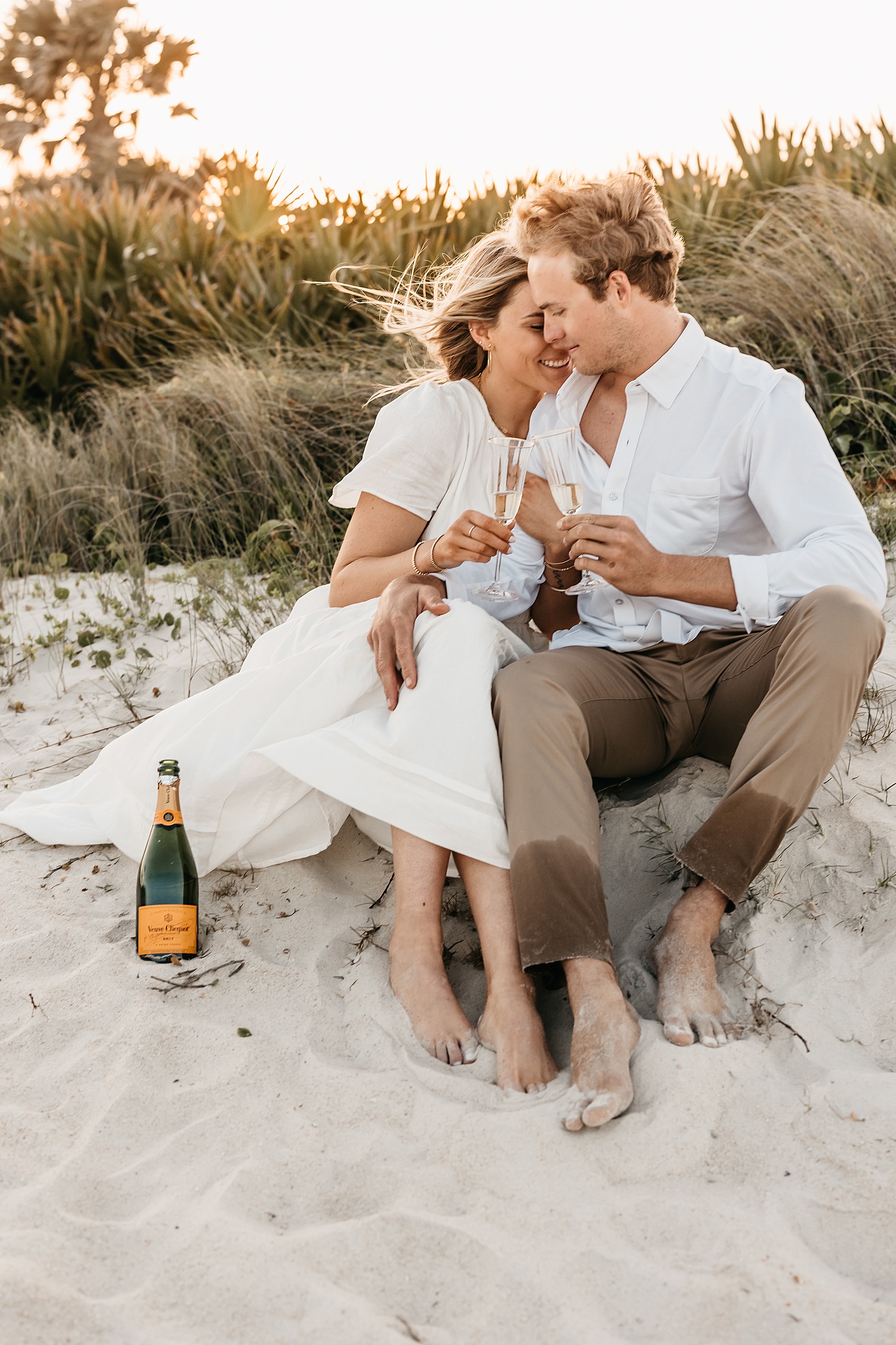 man and woman sitting on the sand beach holding champagne