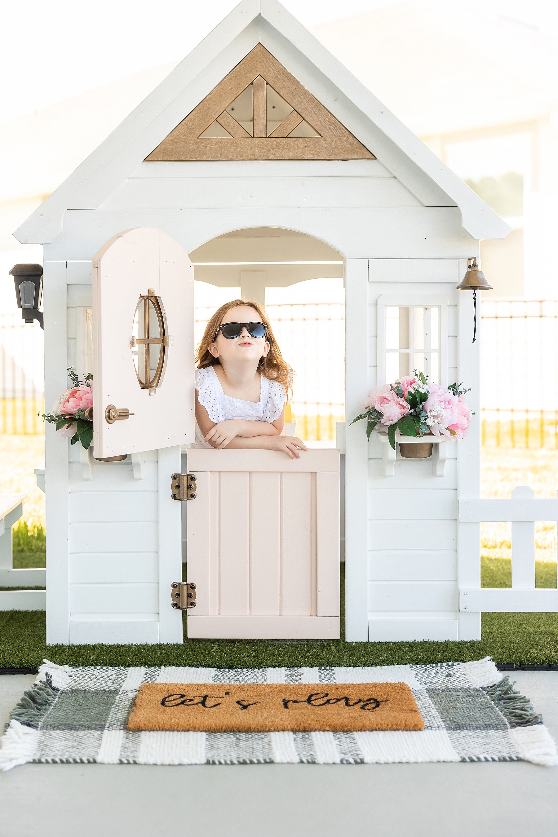 white kids playhouse with pink door and pink and white flowers in pots