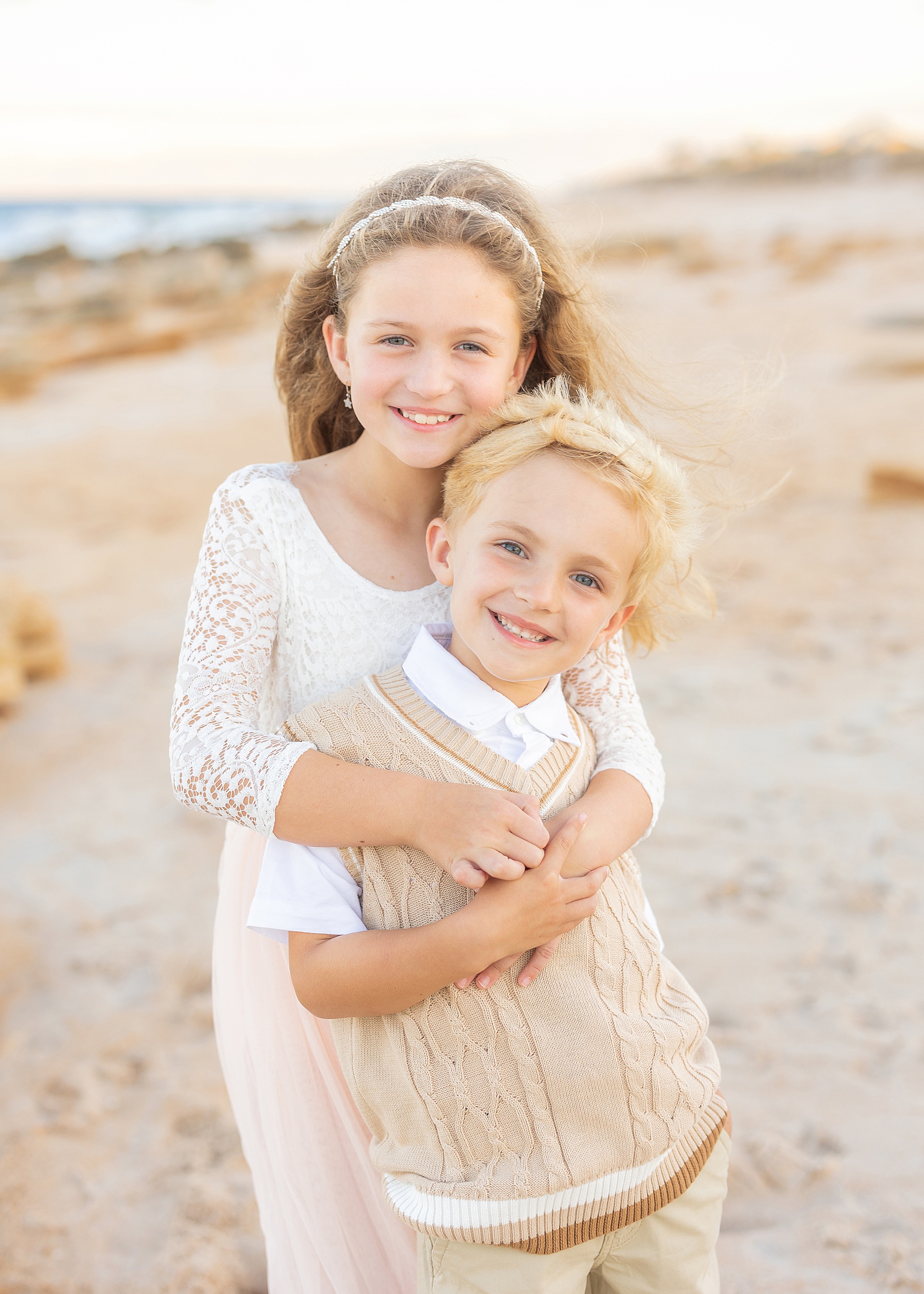 little blond haired boy standing with sibling wearing neutral colors on the beach at sunset