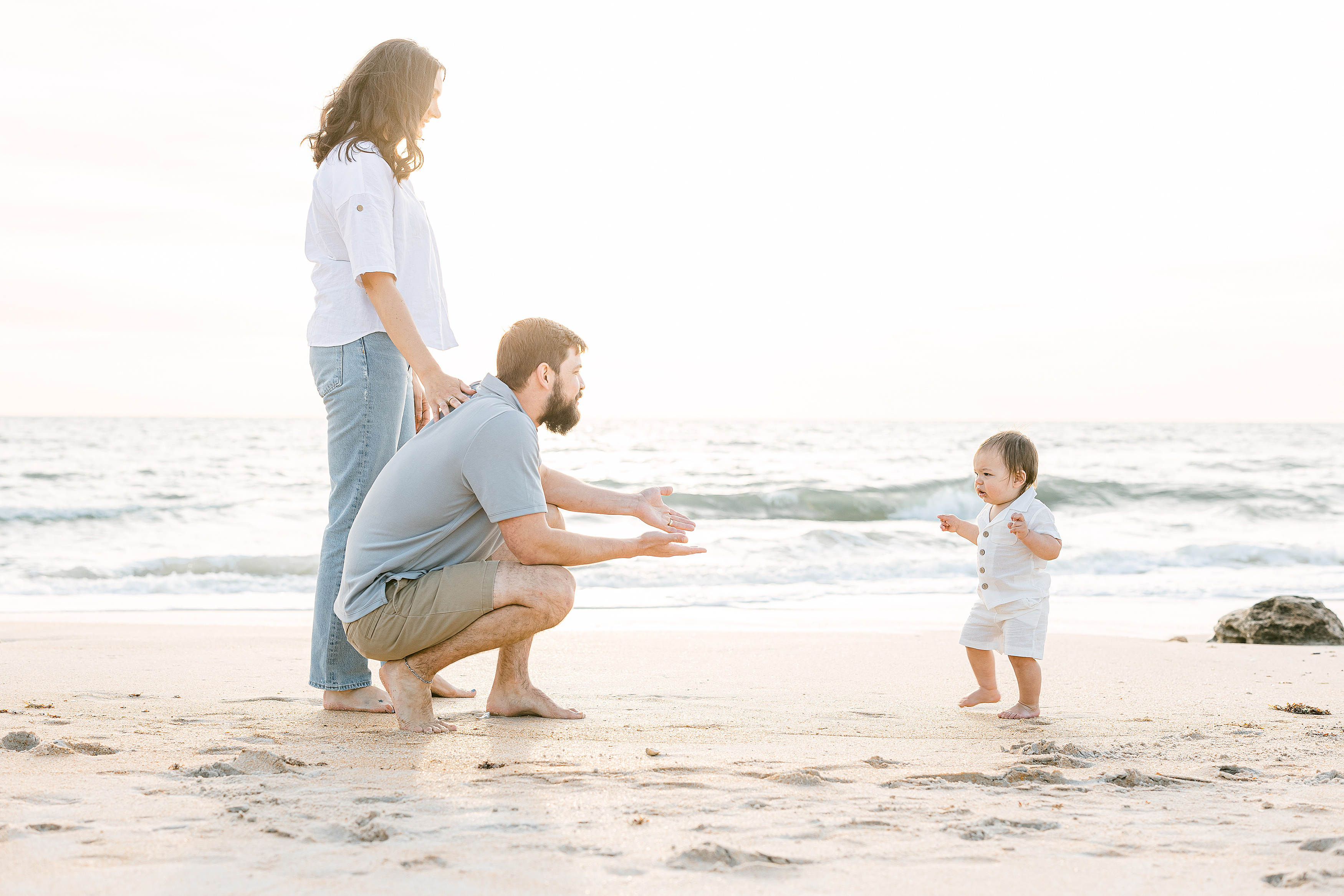 family seeing baby boy walk on the beach at sunrise