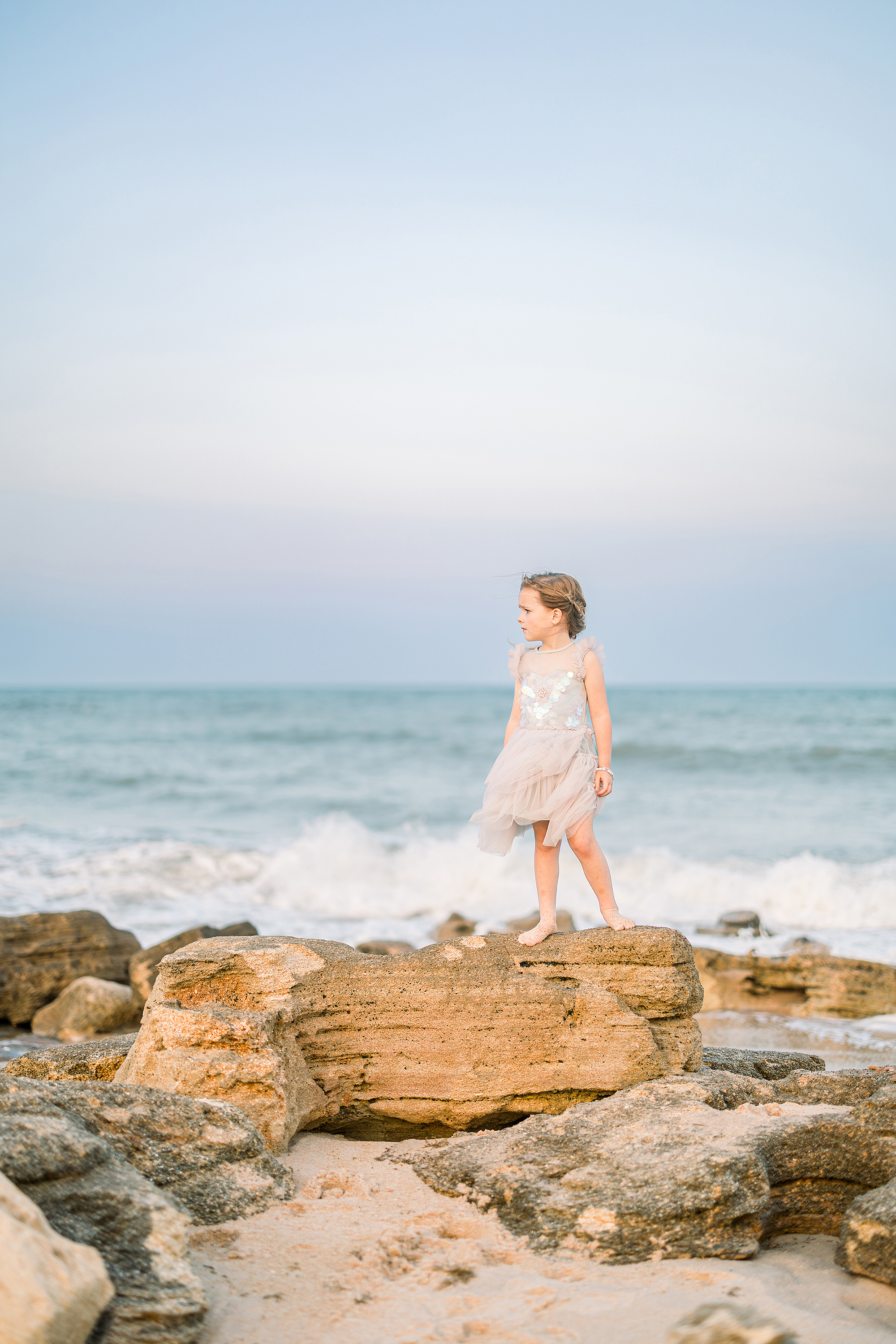 little girl in lilac tutu dress standing on the rocks at the beach at sunset