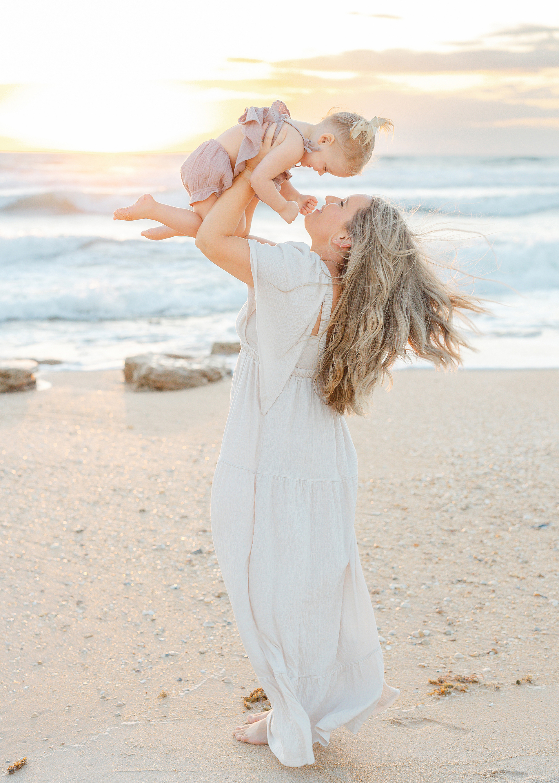 Woman holding baby girl in the air on the beach at sunrise kissing.