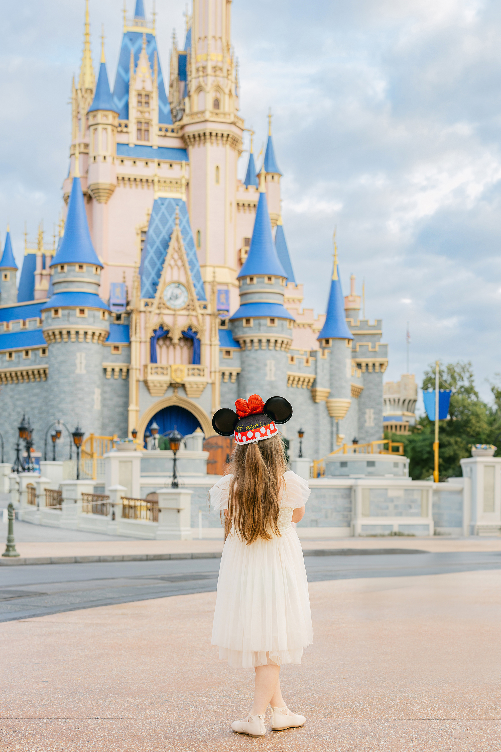 A little girl dressed in a cream tulle dress wears a Mickey hat and looks out at Cinderella's Castle.