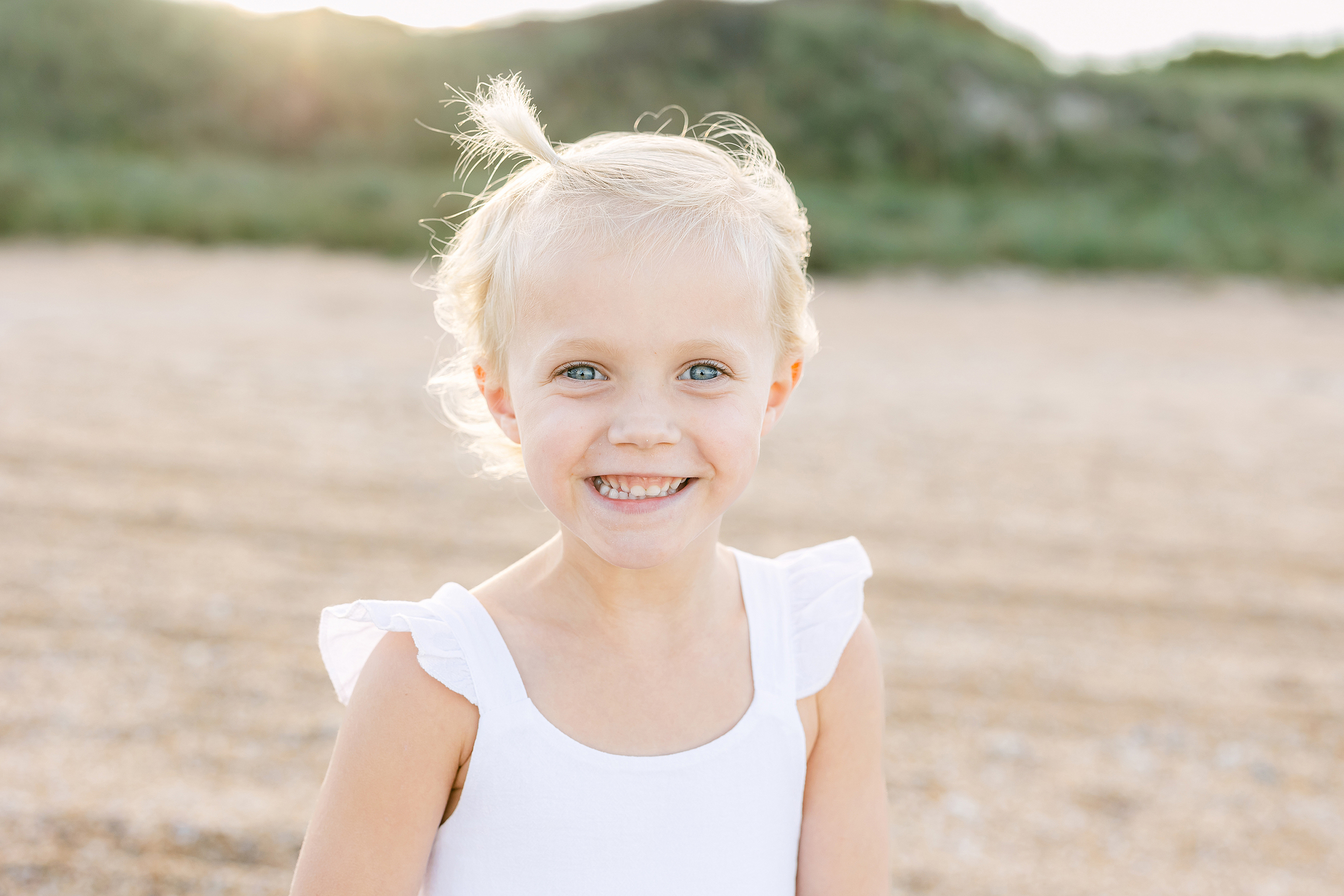 Little girl with blonde hair and blue and white dress smiling on the beach.