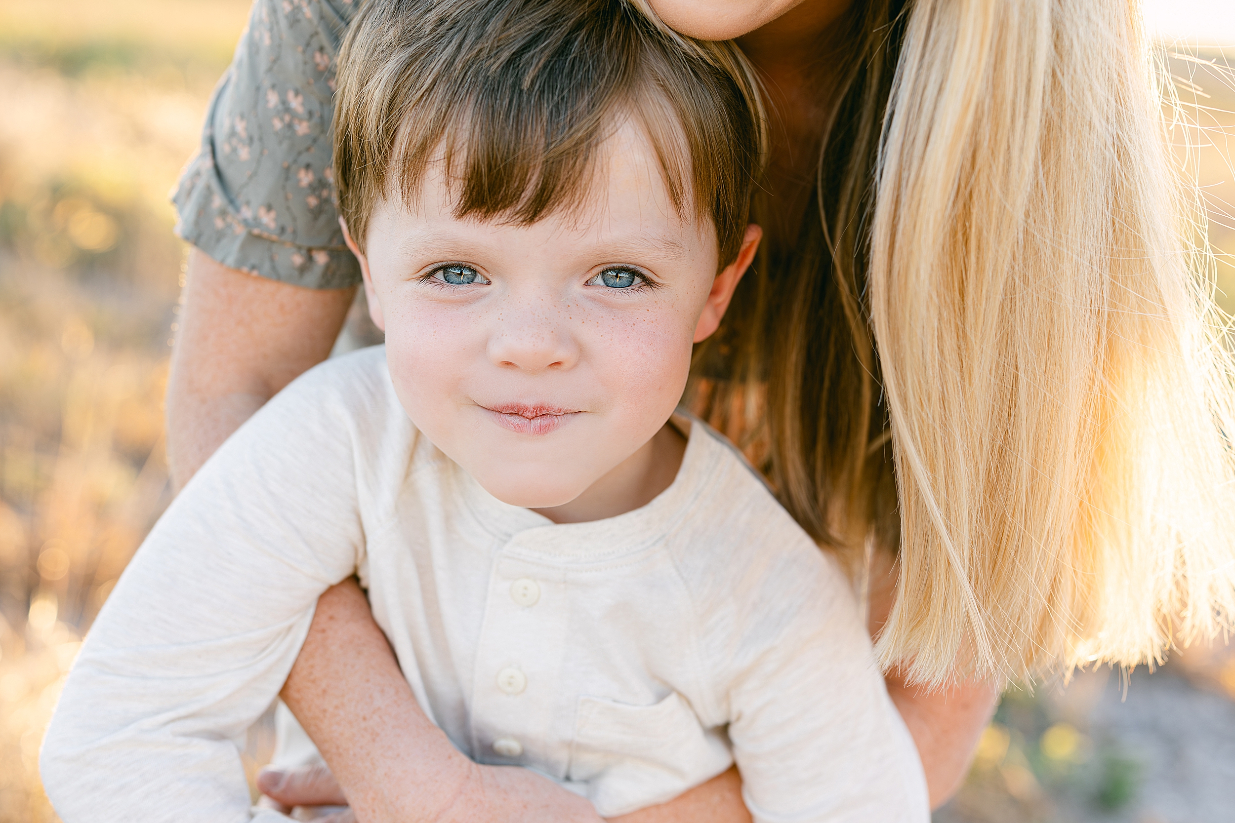 A portrait of a little boy wearing a white long-sleeve henley stares at the camera with blue eyes.