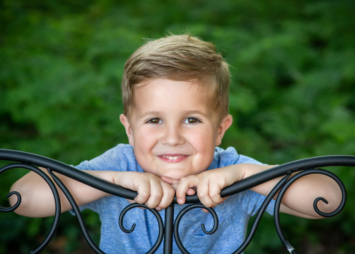 Smiling child in a blue shirt leaning on decorative black railing, with greenery in the background.