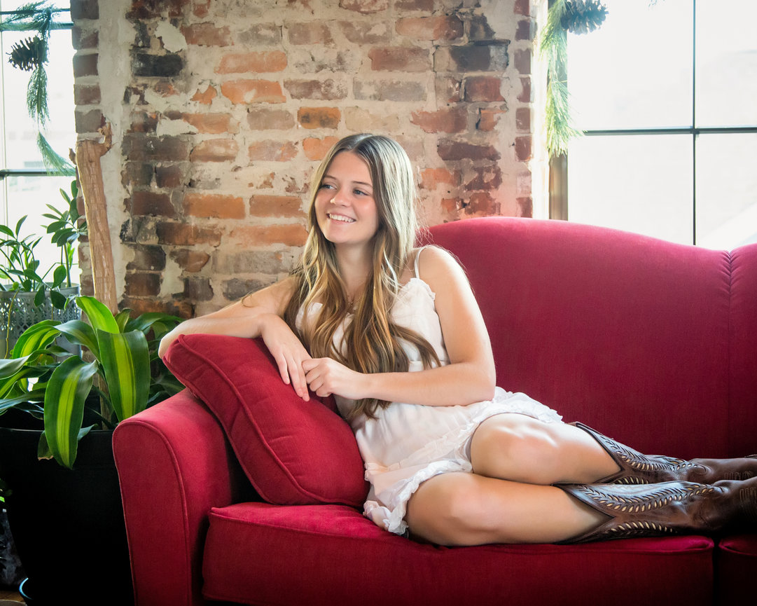 Woman in white dress and boots lounging on red sofa near brick wall.