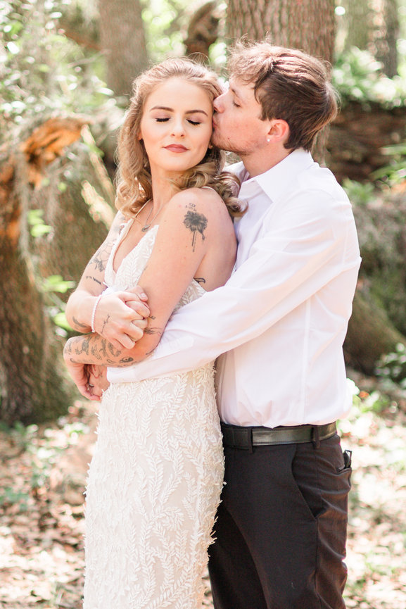 Couple embracing in a forest at Mill Pond Estate. Groom kissing bride's temple. Bride in lace dress, groom in white shirt and dark pants.