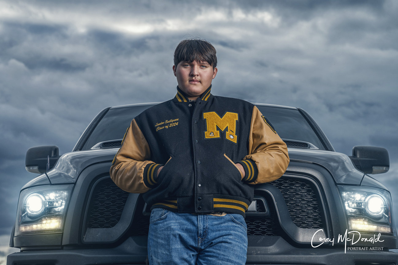 Teen in letterman jacket stands confidently in front of a truck under a cloudy sky.