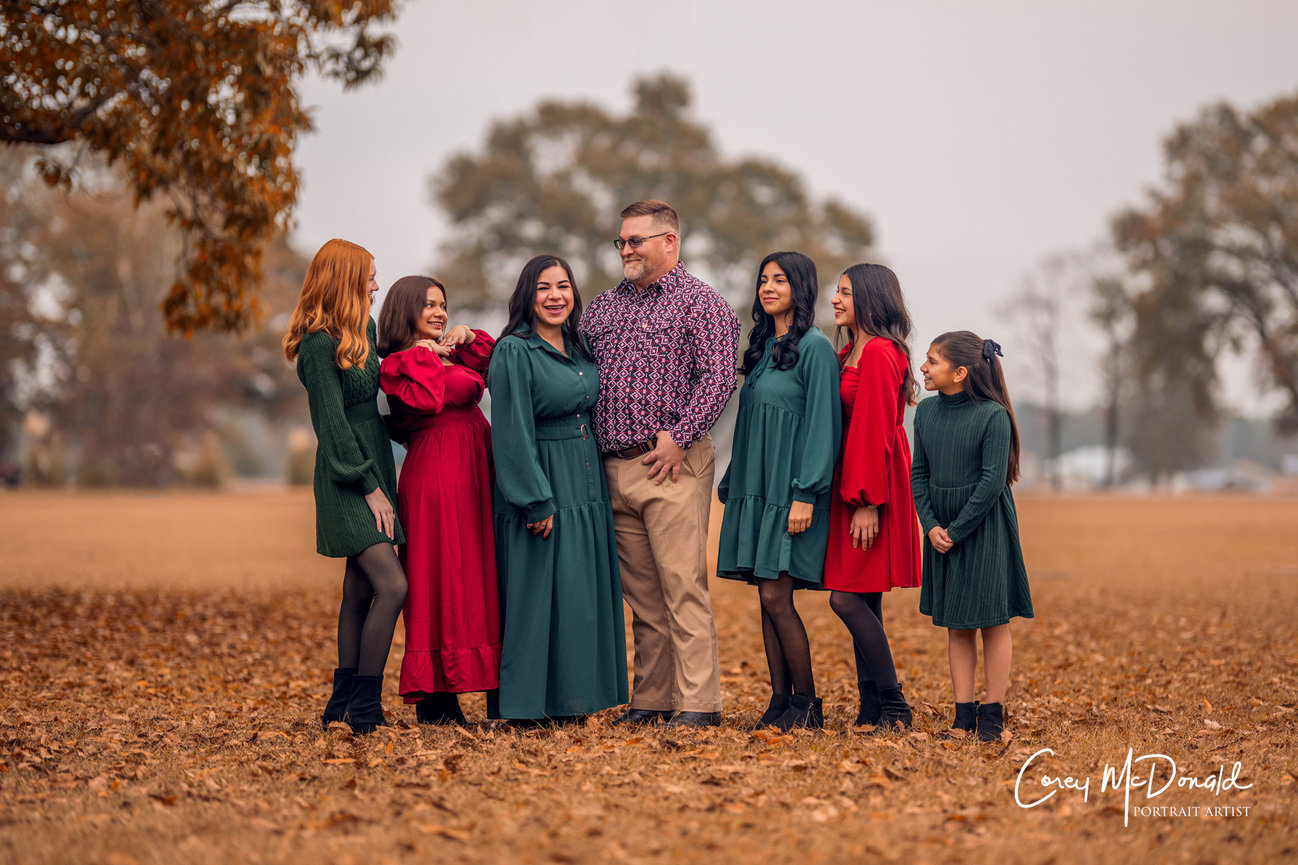 A group of seven people, wearing red and green outfits, stand together outdoors on a leaf-covered ground.