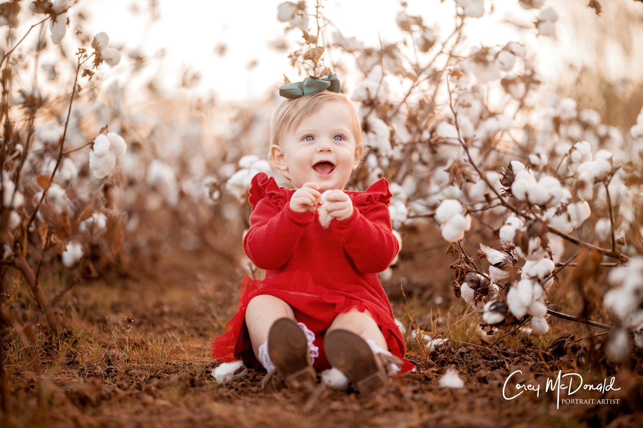 Baby in a red dress sitting among cotton plants, smiling with a green bow in hair.