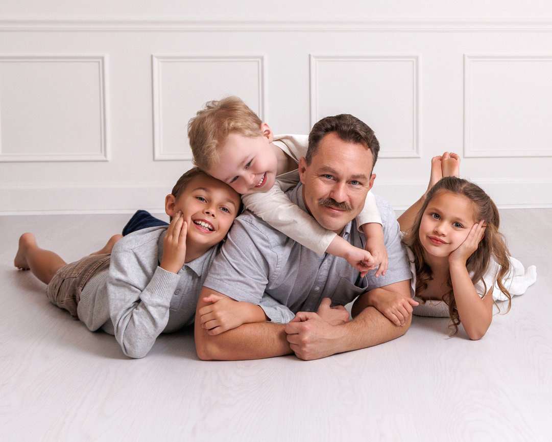 Father and three children lying on the floor, smiling in a cozy, white-paneled room.
