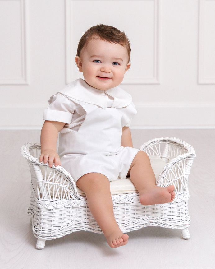 Baby wearing a white outfit sitting on a small white wicker chair, smiling in a minimalist room.