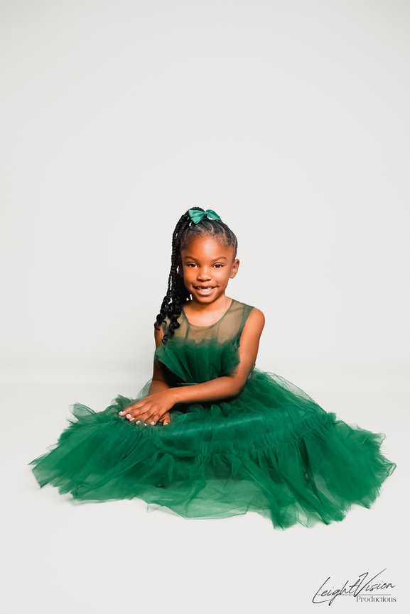 A young girl in a vibrant green dress poses for a photograph, radiating happiness and charm in her expression.