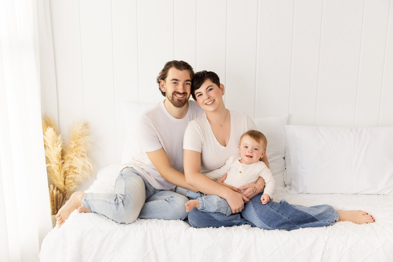 Parents seated with their baby on a white studio bed in soft neutral clothing