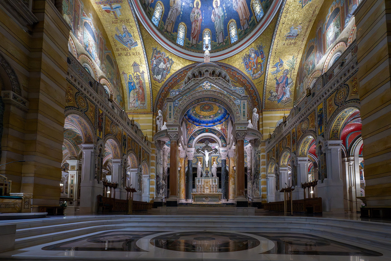 Interior of the Cathedral Basilica of Saint Louis showing the ornate sanctuary, central altar, marble columns, arches, and colorful gold-and-blue mosaics.