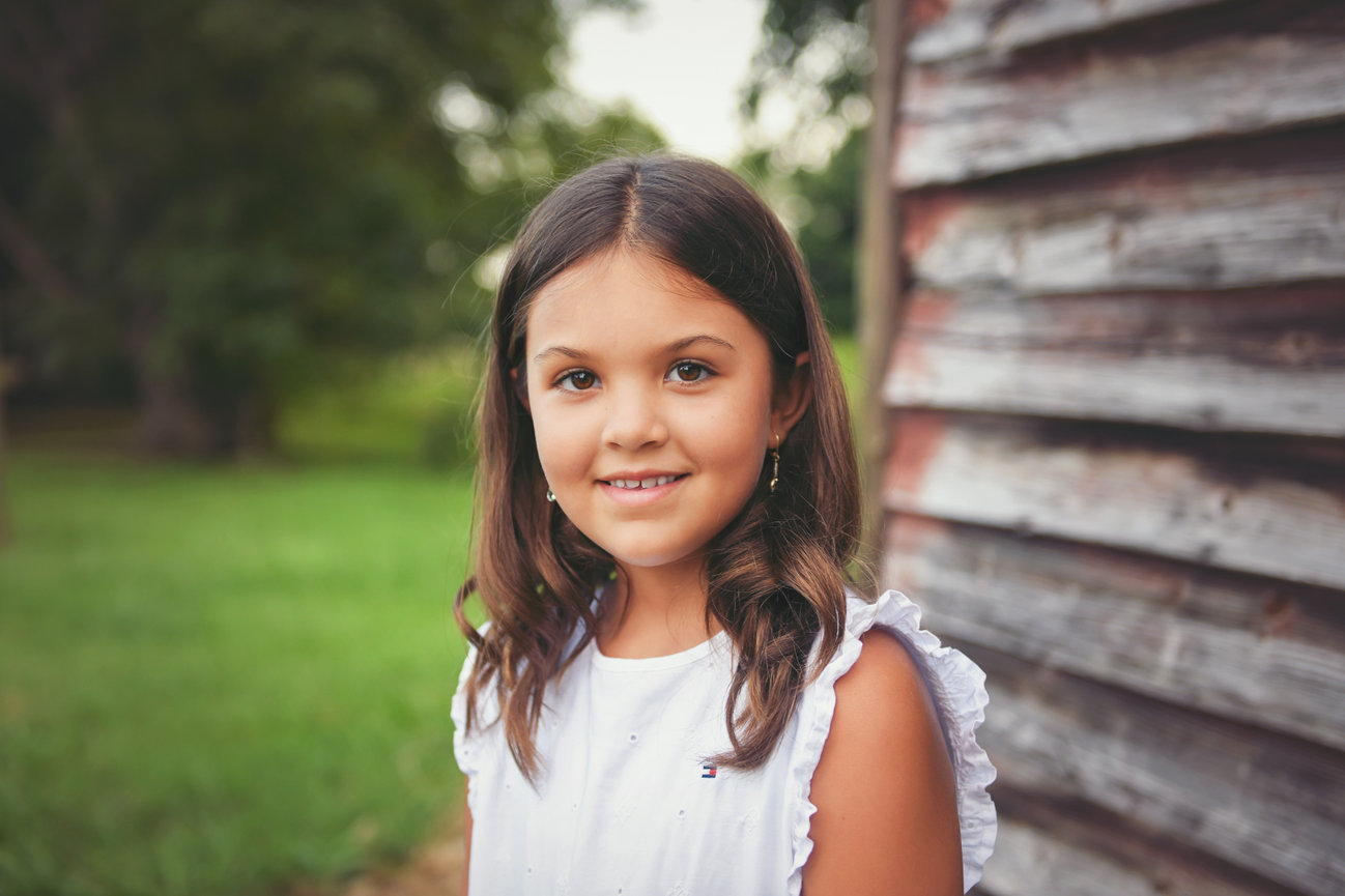 Smiling young girl in a white dress standing next to a weathered barn during a summer child portrait session in Mocksville, NC by M.Gioeli Photography