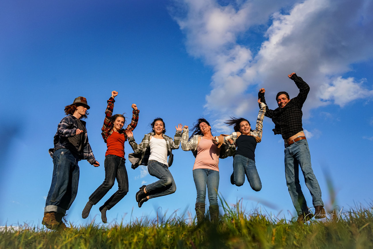 Six people jumping in the air on a grassy field under a clear blue sky with clouds.