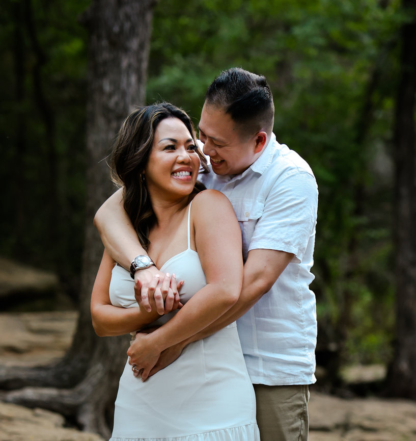 A couple embraces joyfully in a forest, both smiling and dressed in casual summer attire.