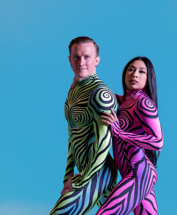 Performers in vibrant body suits posing closely against a blue backdrop
