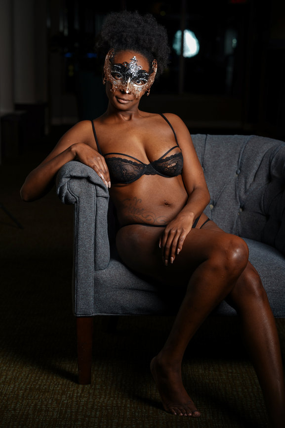 Woman wearing a decorative mask and lingerie, seated on a gray upholstered chair in a dimly lit setting.