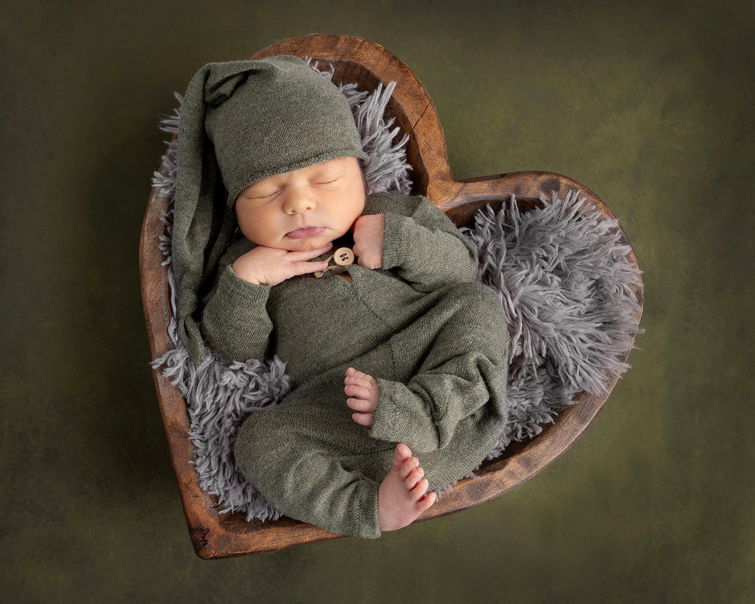 Newborn in green outfit sleeping in a heart-shaped bowl