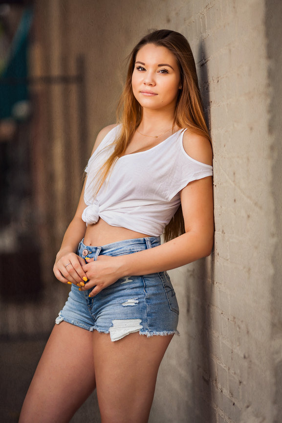 High school senior girl in a white top and denim shorts leaning against a wall by San Antonio photographer Amado Rivas.