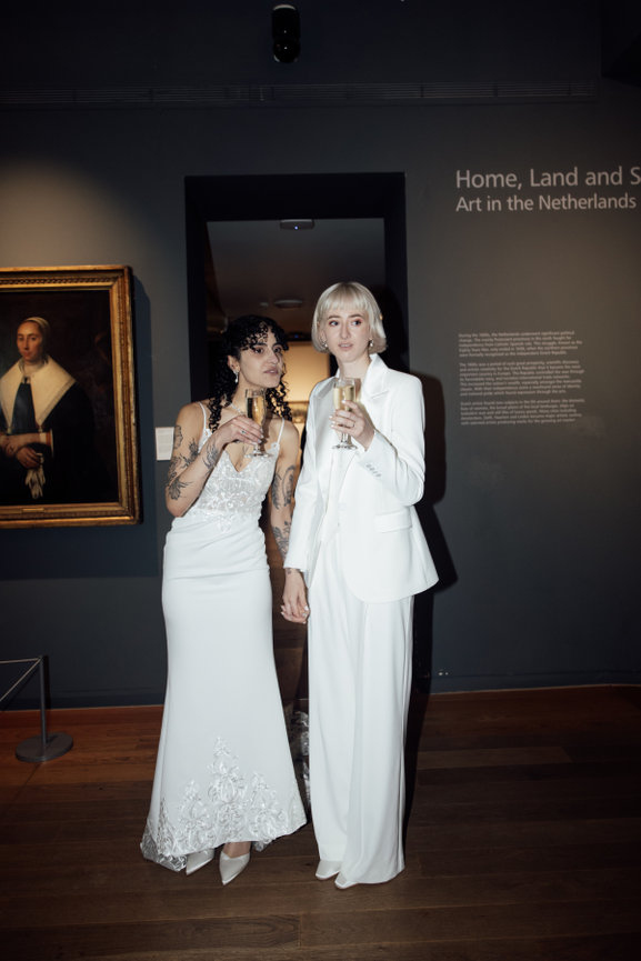 Editorial wedding photography of two brides in elegant white attire drinking champagne, standing by a framed portrait in Manchester Art gallery captured with on-camera flash.