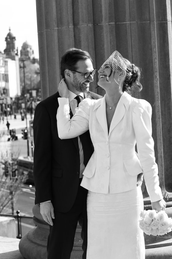 Editorial B&W wedding photography: A smiling couple in formal attire stands outside St Georges Hall, Liverpool near large columns. The woman holds a bouquet and wears a birdcage veil.