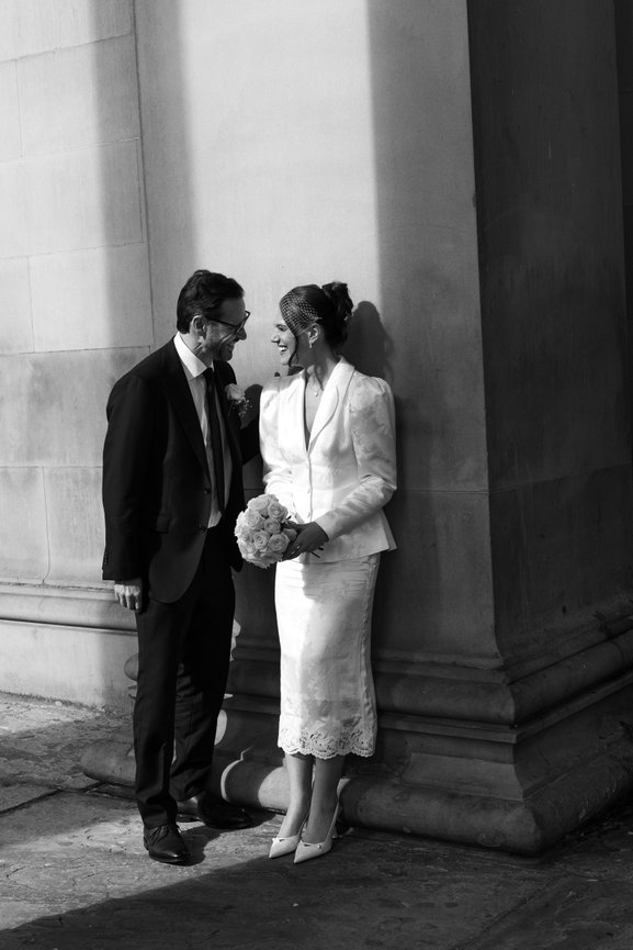 Editorial City Wedding Photography: A couple in formal attire stands against a building's corner, smiling at each other; she holds a bouquet.
