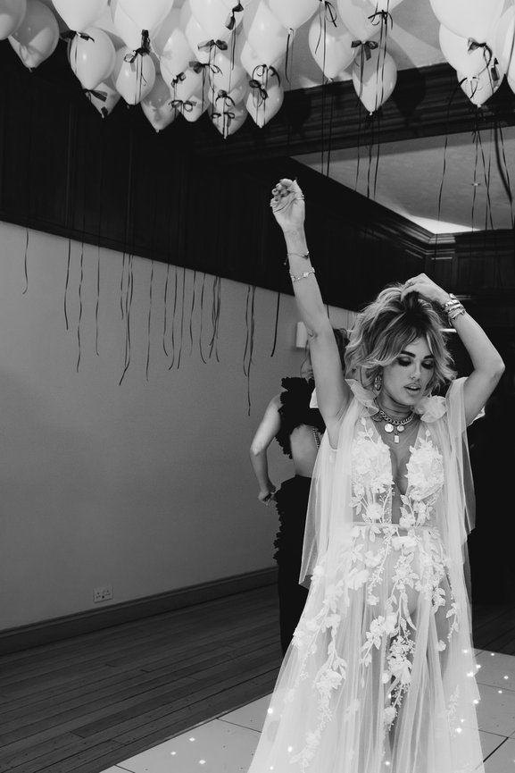 Editorial wedding photography: Bride, Tara Maynard, celebrates wedding anniversary at Standon Hall, the Salt House. Wearing long white wedding dress with 3D Floral Appliqués, balloons fill ceiling of dance floor, captured in black and white.