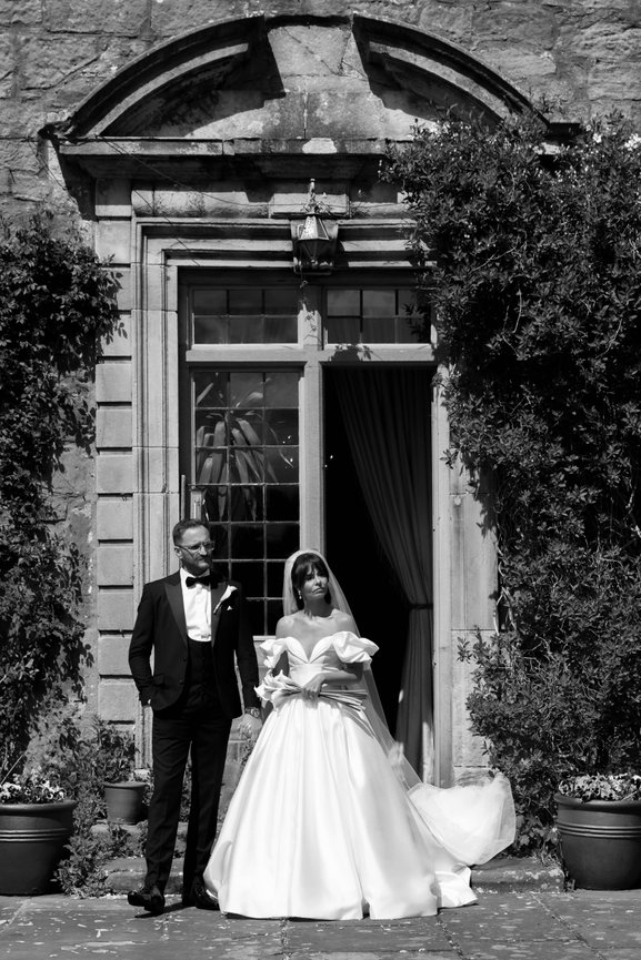 DOCUMENTARY WEDDING PHOTOGRAPHY: Candid moment caught in black and white as bride, holds white lily bouquet, and groom in black tie, hold hands waiting for their confetti toss outside rear entrance of Askham Hall.