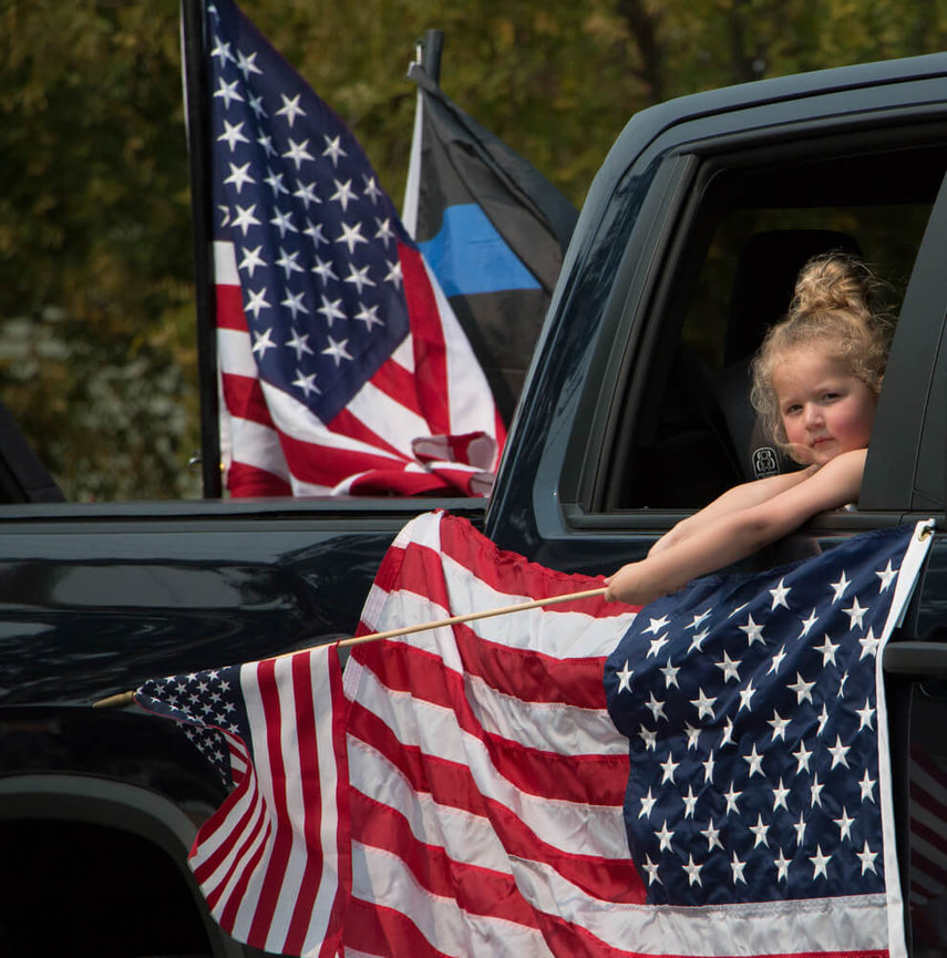 Child in a vehicle holds American flag during a parade, surrounded by other American flags and trees in the background.
