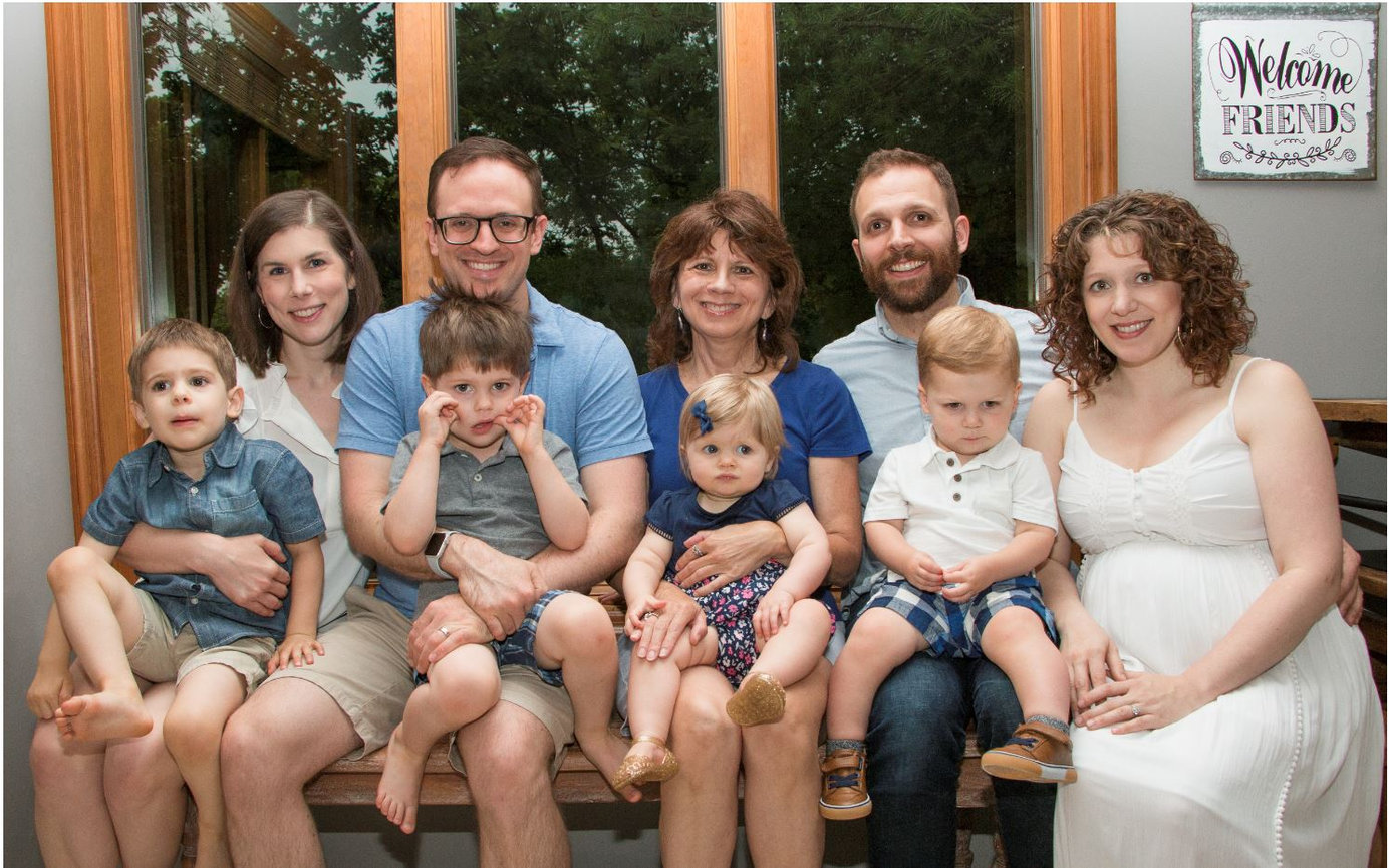 Family smiling, sitting on a bench indoors; includes four adults and four young children. Sign in background says Welcome Friends.
