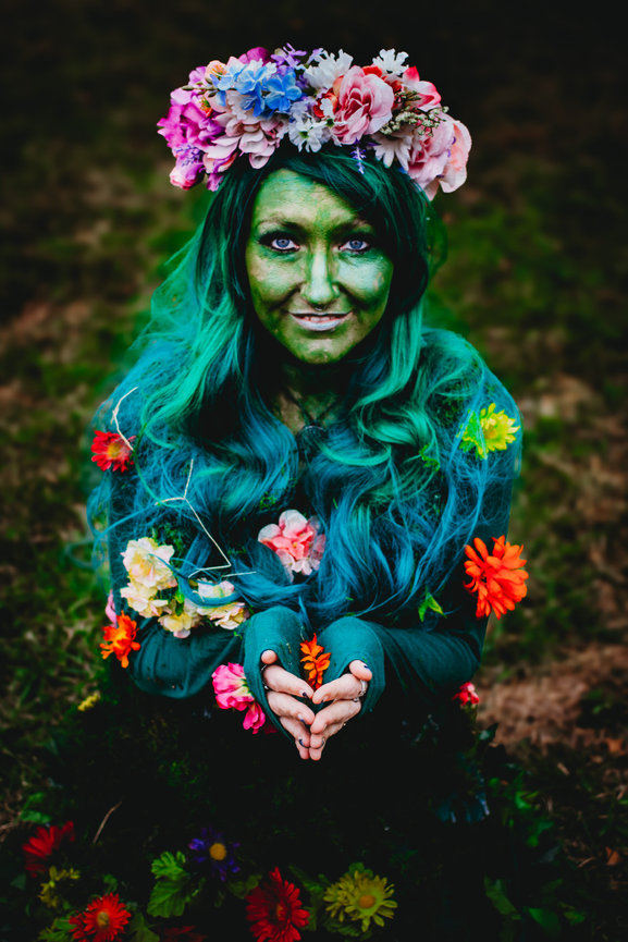 Person with green skin and blue hair, adorned with flowers, crouching and holding a small orange flower in hands.