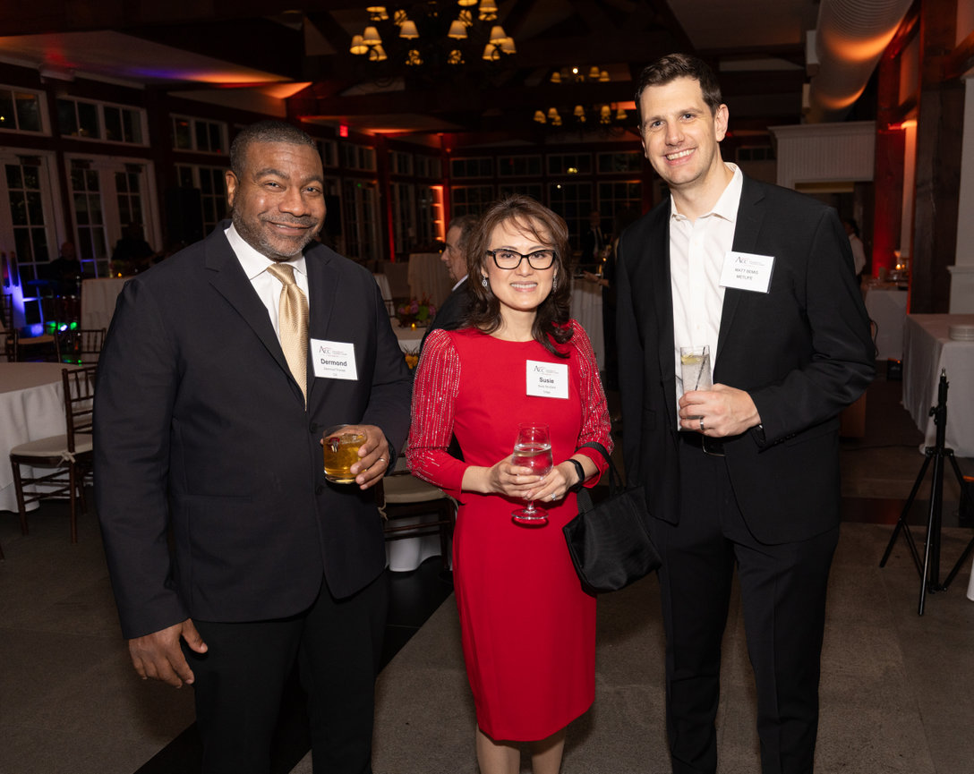 Three people in formal attire at an indoor event holding drinks and wearing name tags.