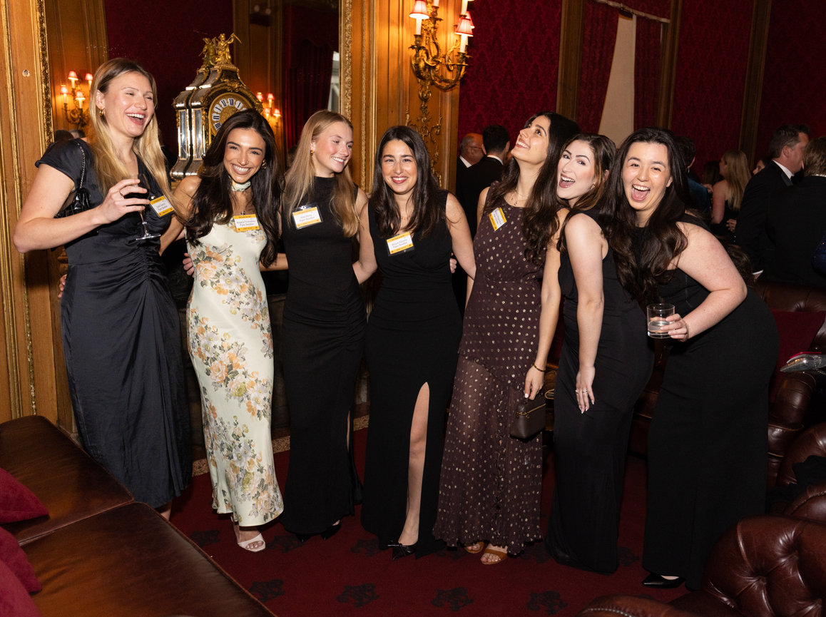 Seven women in elegant evening dresses pose and smile at a formal event in a lavish, warmly lit setting.