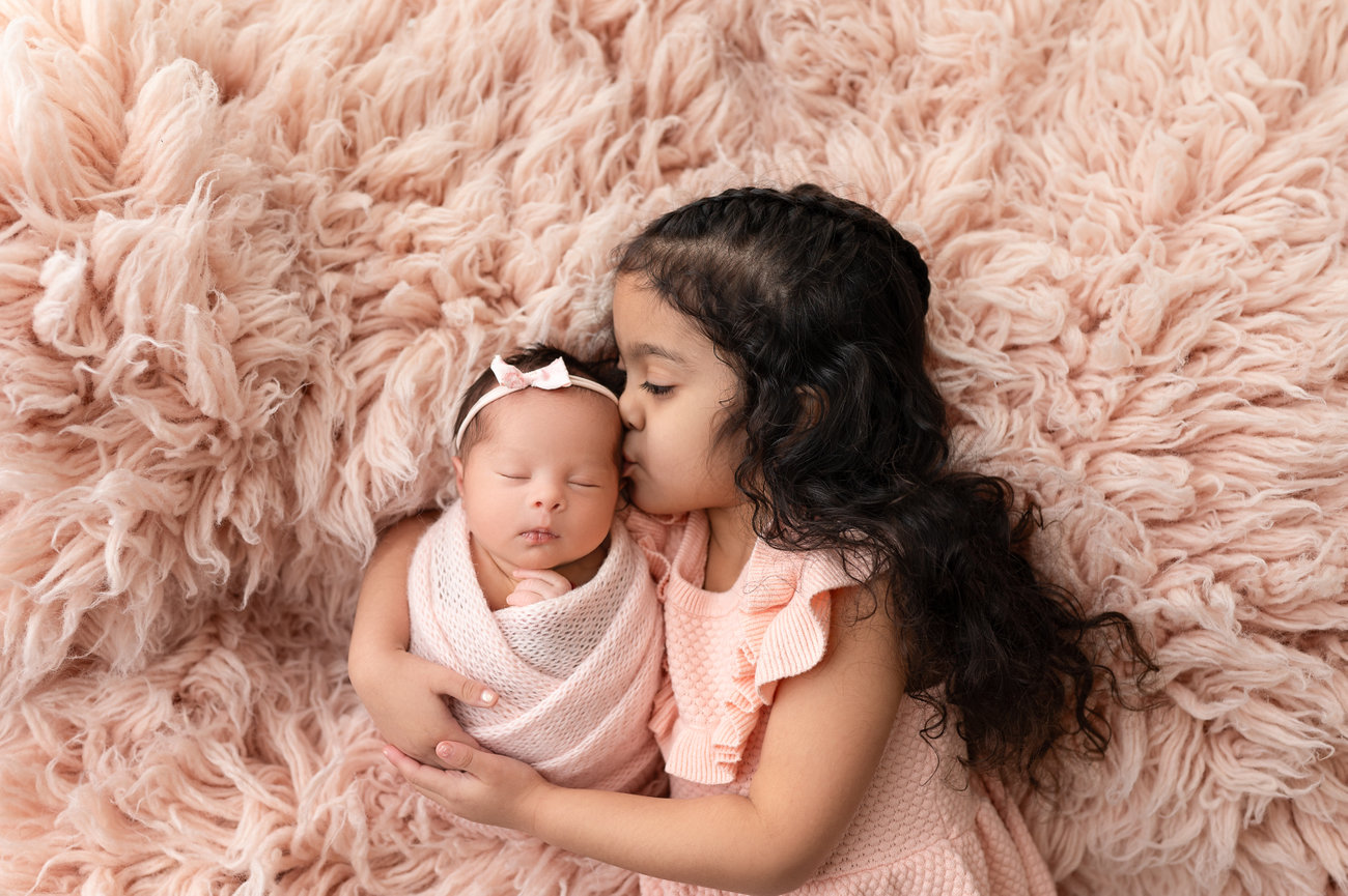 Candid sibling love captured in studio: toddler girl with newborn sibling posed on soft rug backdrop