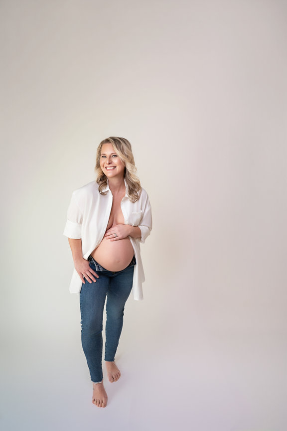 Candid pregnancy picture of mom wearing jeans and an oversized mens dress shirt in a studio setting.