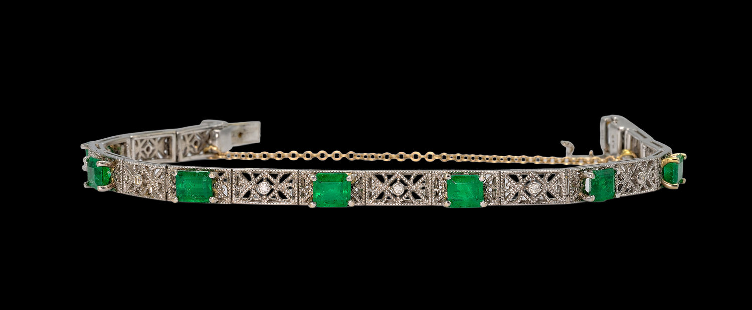 Elegant bracelet featuring square-cut green emeralds set in intricate silver filigree panels with small round diamonds, displayed against a black background.