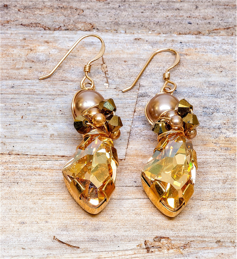 Pair of gold drop earrings featuring large faceted golden crystal pendants topped with gold beads and metallic cube accents, displayed on a rustic wooden surface.