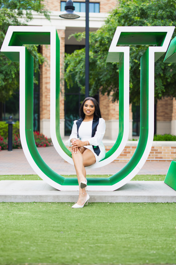 Person sitting on a large green U sculpture at the University of North Texas campus, surrounded by greenery and buildings.