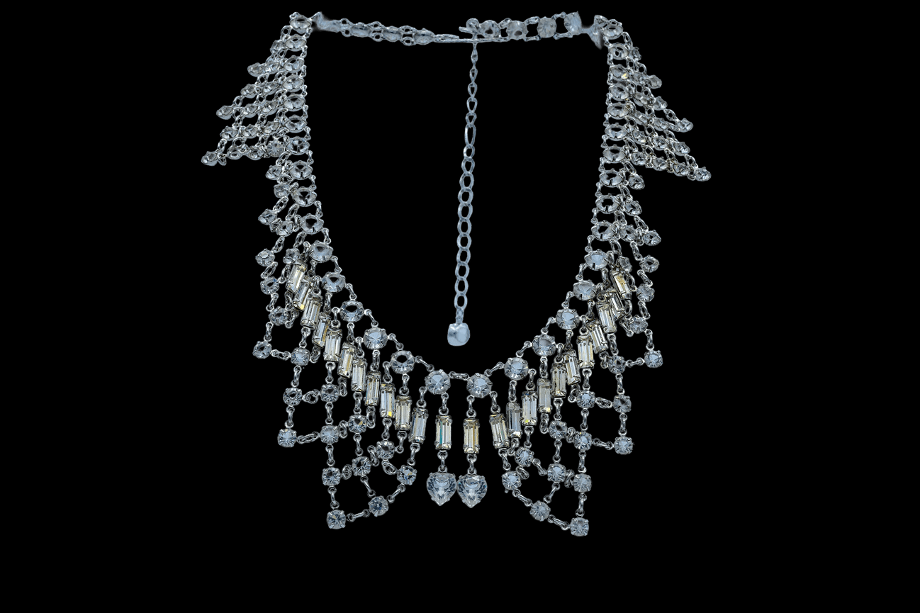 An ornate silver statement necklace featuring cascading rows of round and baguette-cut clear gemstones. The intricate layered design creates a draped effect, accented with two heart-shaped stones at the center, displayed against a black background.