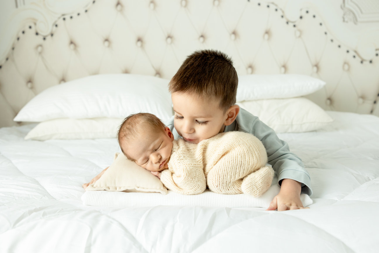 Young child cuddles a sleeping baby on a white bed, both in cozy outfits.