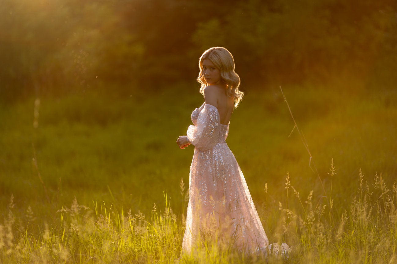 Woman in a sparkling dress standing in a sunlit grassy field, gazing over her shoulder.