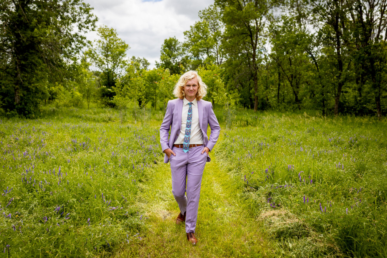 Person walking in a grassy field wearing a light purple suit and colorful tie, surrounded by trees and a cloudy sky.