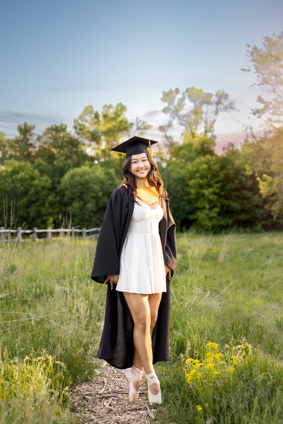 Smiling graduate in cap and gown standing on a grassy path at sunset