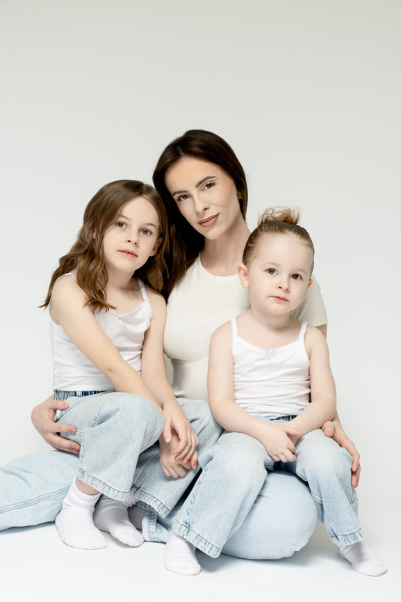 Mother with two young daughters sitting together in white tops and light jeans on a white background