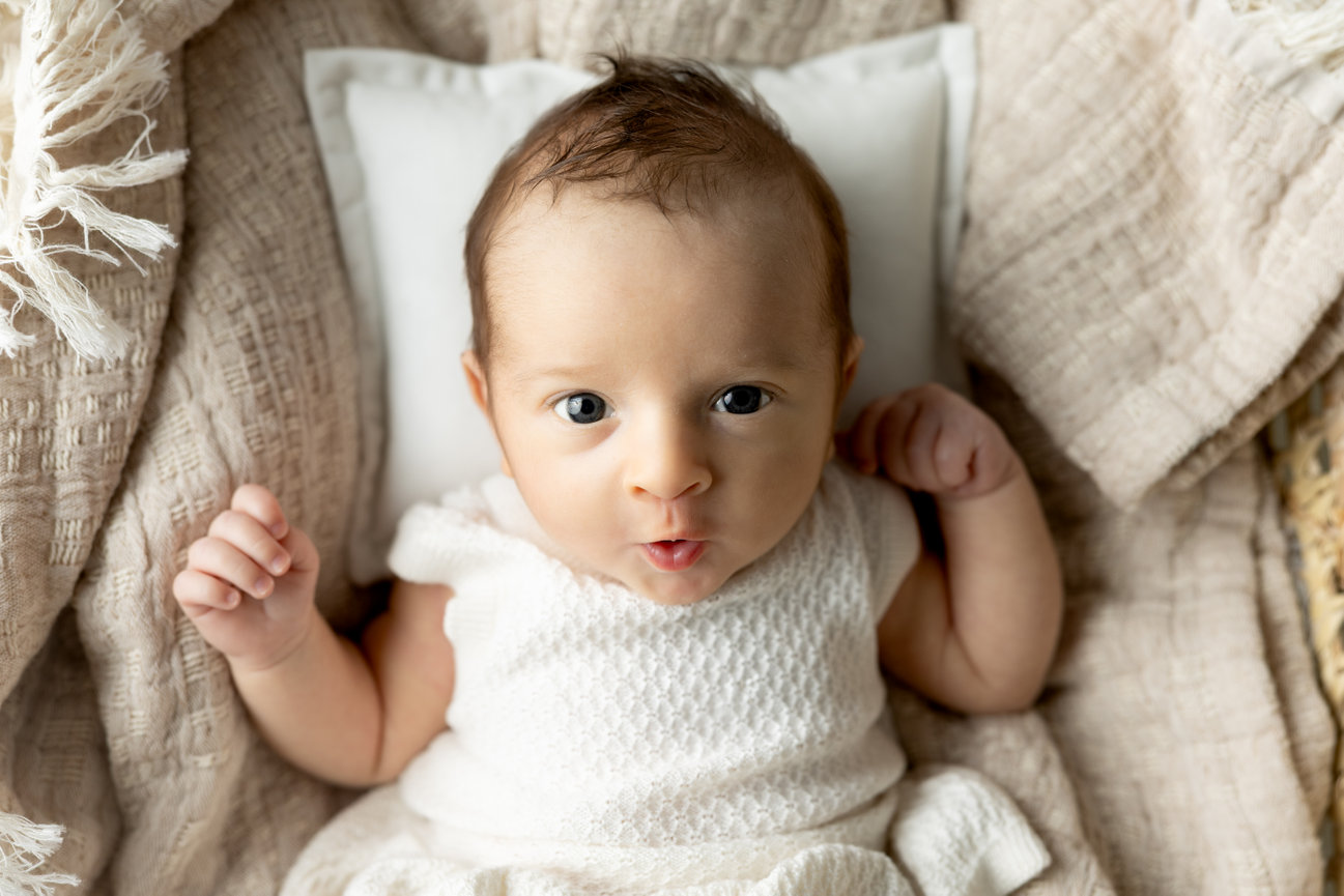 Newborn baby in a white outfit lying on a beige blanket, looking up with wide eyes.