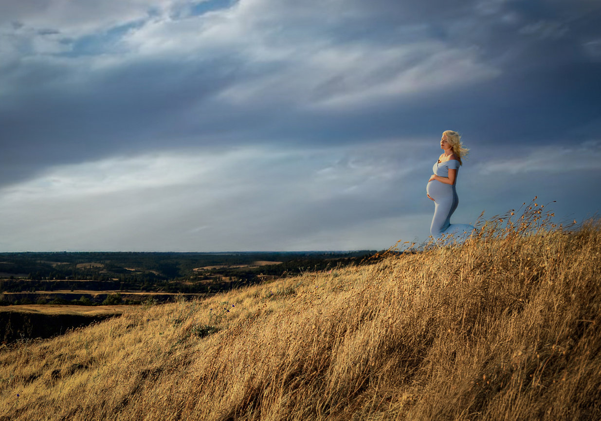 Woman standing on a dry grassy cliff overlooking a wide landscape.